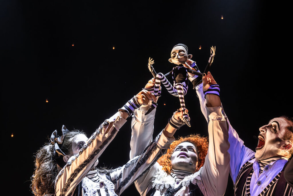Um boneco com uma roupa listrada é segurado no alto por três artistas, um homem e duas mulheres, em trajes teatrais e maquiagem preta e branca que destaca os ossos do rosto Os trajes deles apesar de diversos, são todos numa paleta de cor preta e branca com listras, assim como a roupa do boneco. Cada artista estende a mão em direção ao boneco contra um fundo escuro com luzes sutis semelhantes a estrelas. Foto: Renato Mangolin