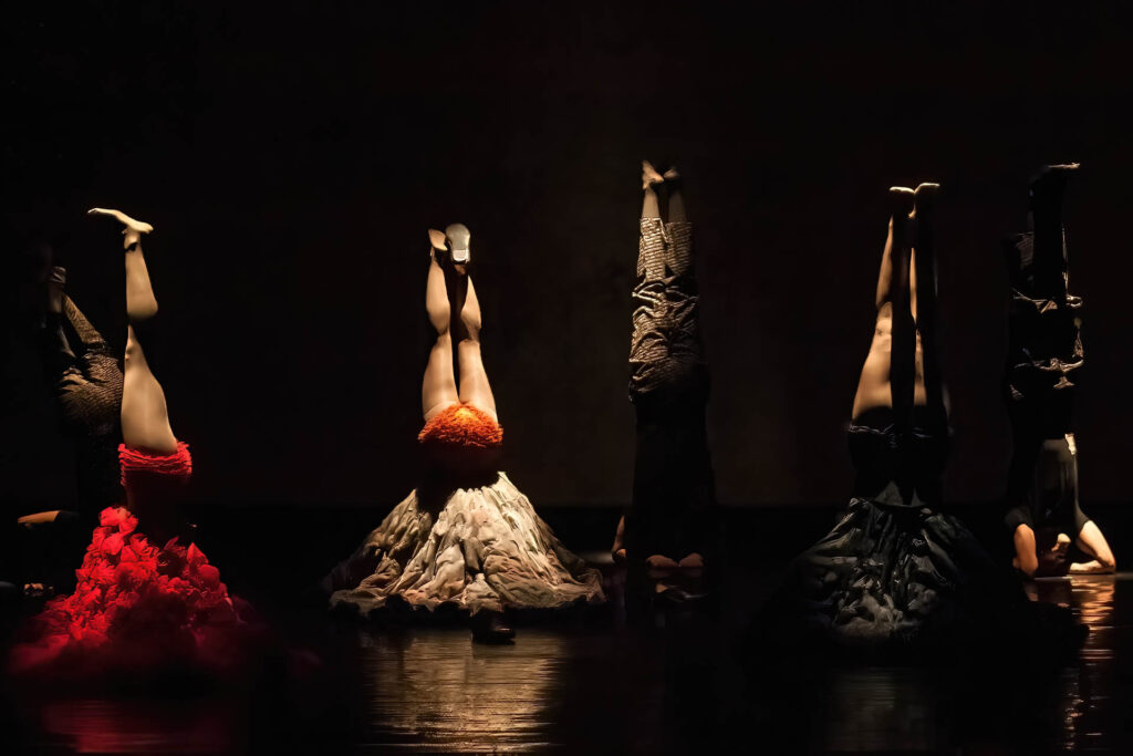 Cinco dançarinos, mulheres e homens, fazem parada de cabeça no palco com pouca iluminação, alguns usando saias texturizadas em camadas em várias cores e padrões e outros com calças escuras. O holofote dramático destaca os designs exclusivos de suas saias contra um fundo escuro. Foto: Guto Muniz