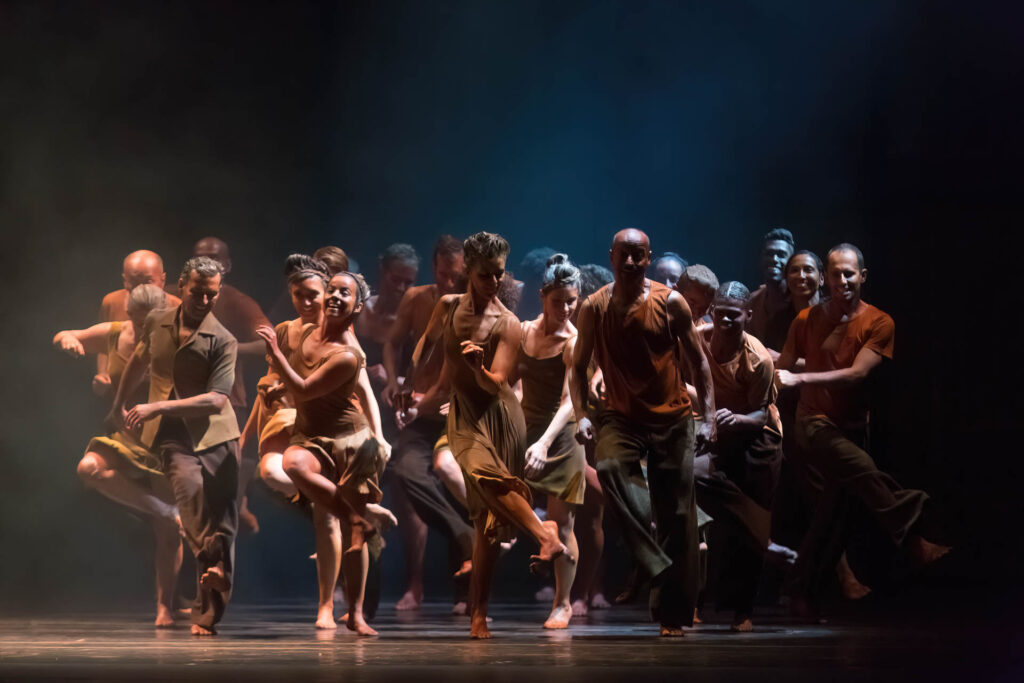 Um grupo de dançarinos e dançarinas se apresentando energicamente no palco. Eles estão usando trajes em tons de terra, e a iluminação cria uma atmosfera dramática com tons escuros e melancólicos. Os dançarinos estão se movendo em uníssono com poses dinâmicas e expressivas. Duas dançarinas no palco fazem uma pose dramática. A dançarina da esquerda está de cabeça para baixo, usando uma saia vermelha que cobre seu tronco de cabeça pra baixo, parecida com uma rosa. O dançarino da direita se inclina para frente em uma roupa preta e dourada. O fundo é escuro e somente as dançarinas são sutilmente iluminadas. Foto: Guto Muniz