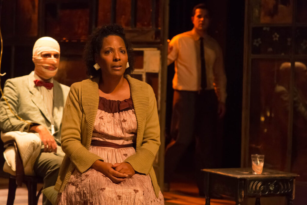 Uma mulher de vestido e cardigan está sentada, parecendo preocupada. Atrás dela, uma pessoa com o rosto enfaixado está sentada em uma cadeira, vestindo um terno e gravata borboleta vermelha. Outra pessoa está de pé ao fundo, como se tivesse saindo por uma porta, de terno e gravata. O fundo parece de madeira com algumas texturas e relevos. A iluminação é quente e suave. Foto: Guto Muniz