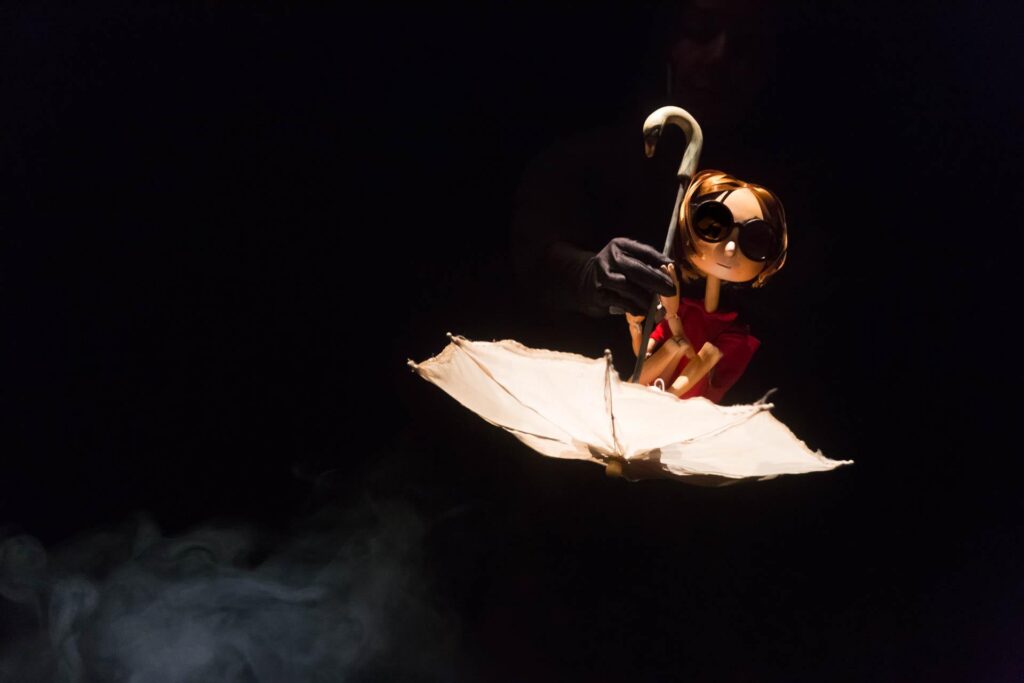 Uma boneca de madeira com cabelo curto e óculos redondos está sentada dentro de um guarda-chuva branco aberto. Uma mão usando uma luva preta segura o pequeno guarda-chuva por trás. A cena é suavemente iluminada contra um fundo escuro. Foto: Guto Muniz