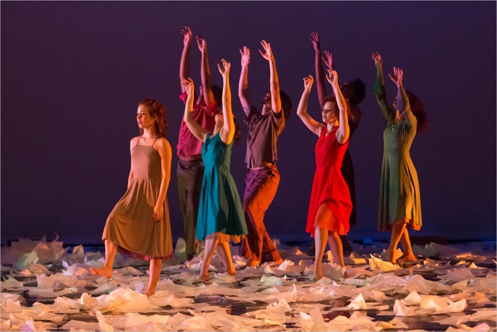 Dançarinos, homens e mulheres, em vestidos e roupas coloridas se apresentam expressivamente com os braços erguidos em um palco coberto de papel branco espalhado. A frente do grupo uma mulher, de vestido marrom claro, tem os braços abaixados junto ao corpo enquanto executa o movimento de caminhar com uma das pernas ainda no ar com olhar distante O fundo pouco iluminado destaca os movimentos vibrantes e poses dinâmicas do grupo, iluminados por luzes suaves e quentes. Foto: Guto Muniz