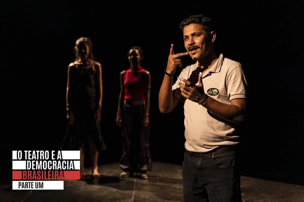 Um homem de camisa bege gesticula expressivamente no palco, com duas mulheres de pé ao fundo, sob iluminação fraca. Foto: Renato Mangolin