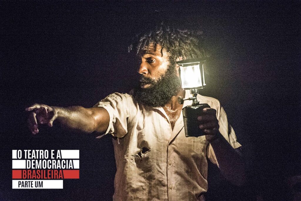 Um homem negro com barba segura uma lanterna, iluminando seu rosto e parte superior do corpo na escuridão. Ele está vestindo uma camisa de cor clara com pequenos rasgos, e aponta para a distância com a outra mão. Foto: Rodrigo Sambaqui