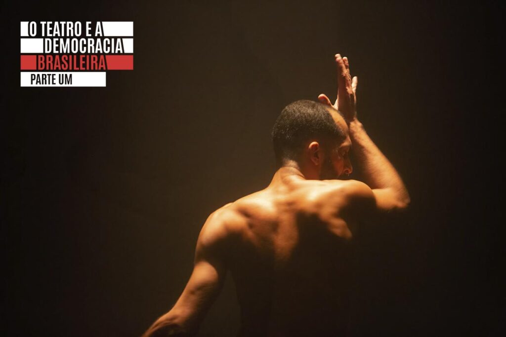 Um homem sem camisa com músculos tonificados está em pé sob iluminação quente e suave em tronco. Ele se encontra de costas, com uma mão tocando a testa. A cena é pouco iluminada, contra um fundo escuro. Foto: Guto Muniz