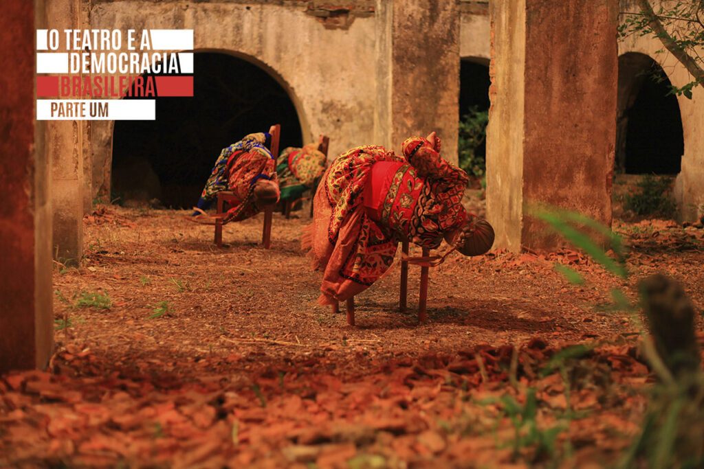 Três mulheres com roupas coloridas se inclinam com seus corpos sobre cadeiras. A cena externa na grama seca e algumas plantas é emoldurada por ruínas. Foto: Pedro Isaías Lucas