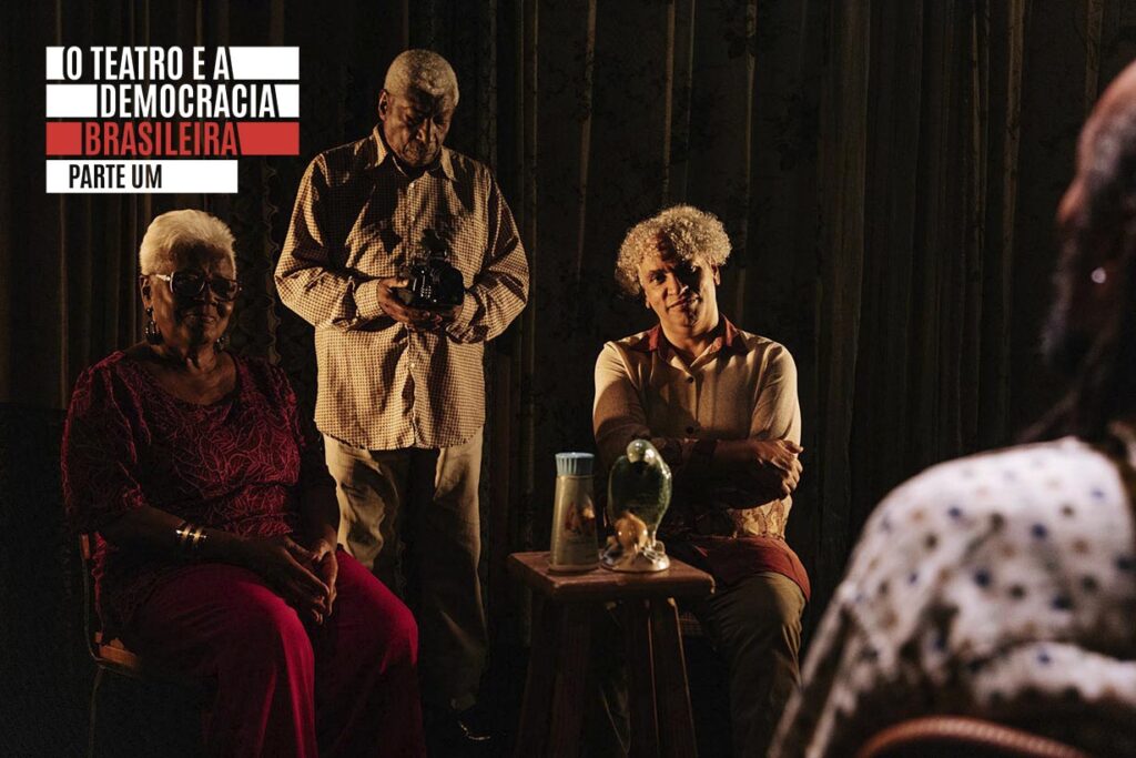 Três idosos negros estão sentados e em pé em um local pouco iluminado. Uma pessoa segura uma câmera, outra está sentada de braços cruzados e uma terceira sorri. Uma pequena mesa com um jarro de vidro e uma garrafa está no centro. no canto direito da foto é possível ver uma quarta pessoa de costas, desfocada, que parece ter características semelhantes e estar olhando para os outros três. A logo do projeto O teatro e a democracia brasileira aparece acima à esquerda. Foto: Noelia Nájera