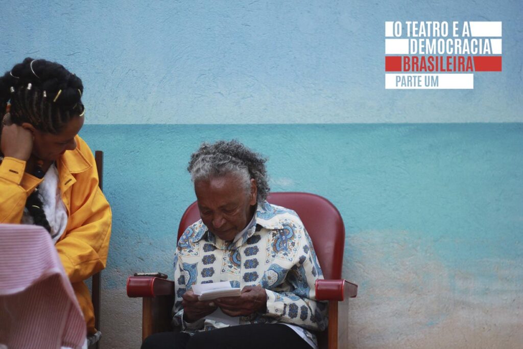 Uma mulher idosa está sentada em uma cadeira vermelha lendo um pedaço de papel, enquanto uma mulher mais jovem ao lado dela, vestida com uma jaqueta amarela, está inclinada com a cabeça apoiada na mão contra uma parede pintada de azul e turquesa. Foto: Tetembua Dandara