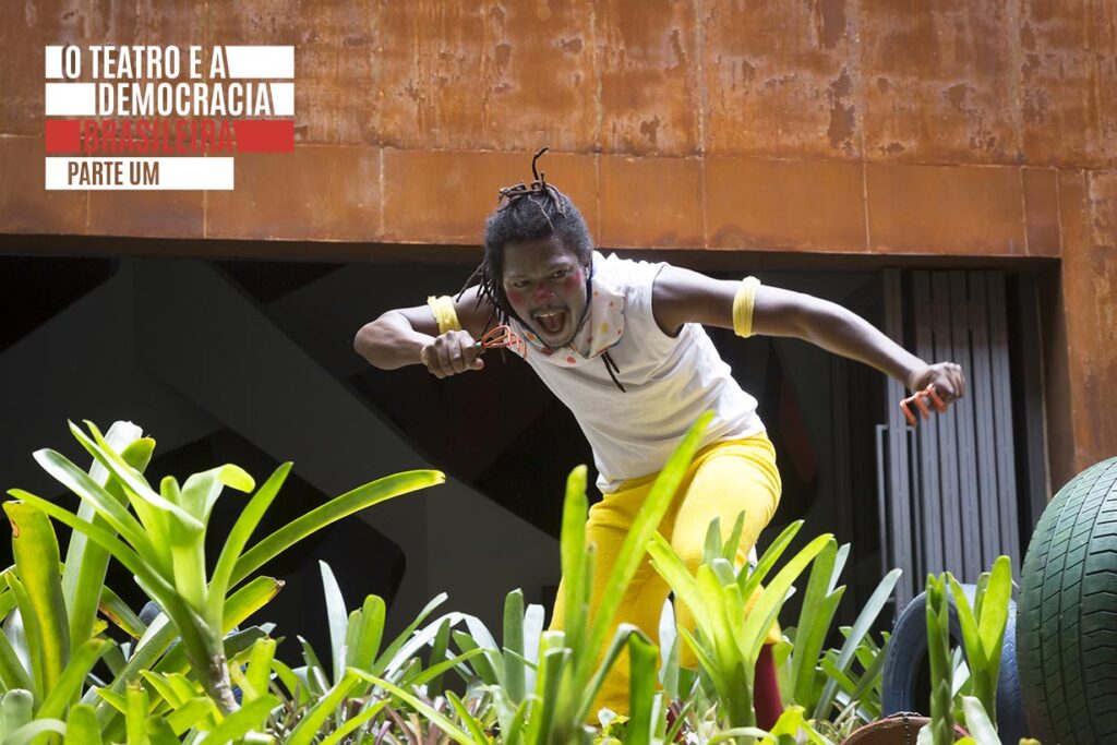 Uma pessoa vestindo camisa branca e calça amarela agacha-se energicamente entre plantas verdes, com um rosto expressivo e punhos erguidos, em frente a uma parede cor de ferrugem. Foto: Guto Muniz